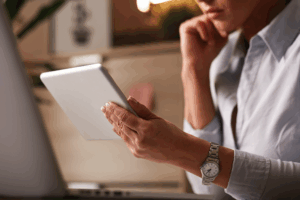 imagem de uma mulher pensativa segurando um ipad ou tablet, na frente da tela de um coputador. Ela veste uma camisa social branca, isa relogio e aparece apenas parte do rosto dela, do nariz para baixo. Ela está com a outra mão apoiada no queixo, pensativa.