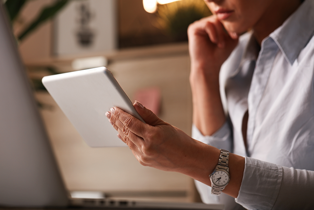 imagem de uma mulher pensativa segurando um ipad ou tablet, na frente da tela de um coputador. Ela veste uma camisa social branca, isa relogio e aparece apenas parte do rosto dela, do nariz para baixo. Ela está com a outra mão apoiada no queixo, pensativa.